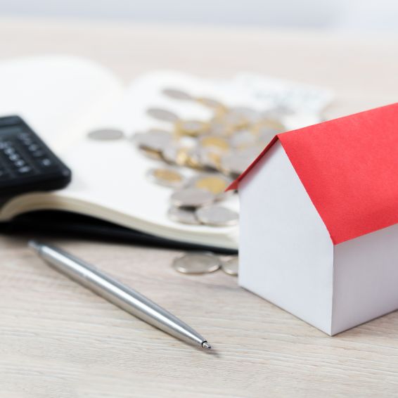 Open notebook with calculator and coins on top next to house-shaped block and a pen