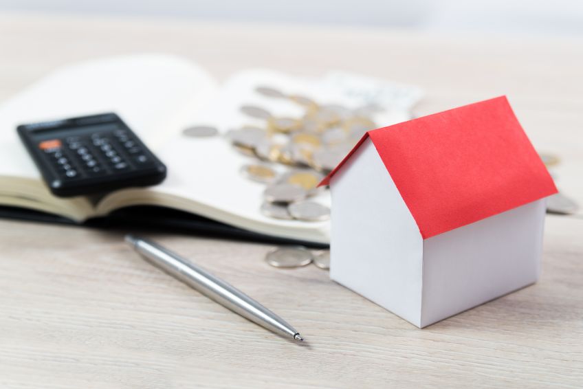 Open notebook with calculator and coins on top next to house-shaped block and a pen